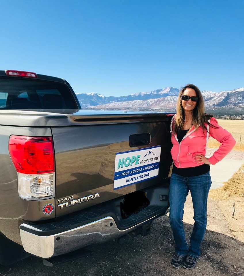 Lori McNamara stands beside her truck with a magnetic sticker that reads, "HOPE IS ON THE WAY TOUR ACROSS AMERICA HOPEPLAYER.ORG'